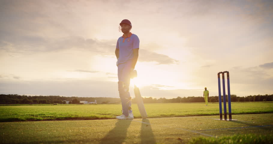 Portrait of a Professional Cricket Player in Blue Uniform and Protective Helmet Waiting for the Game to Start, Standing with a Bat on a Field. Cinematic Low Angle, Warm Sunset Scenery Footage Royalty-Free Stock Footage #1110455127