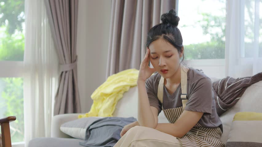 Stressed young woman asian sitting on the sofa in the living room and among a mess at home, woman sitting among scattered clothes on sofa and ignoring, woman frowning and letting out soft sigh.