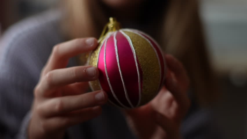 Closeup cropped shot of unrecognizable little girl holding and looking festive Christmas ball. Happy adorable kid decorates Christmas tree for New Year at home in dark living room alone, slow motion.