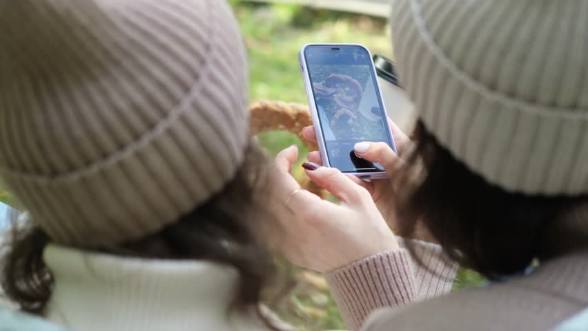 In autumn, two cheerful girls walk through the park during the day, drink coffee and take pictures of glasses of coffee and bagels against the background of autumn foliage for social networks.