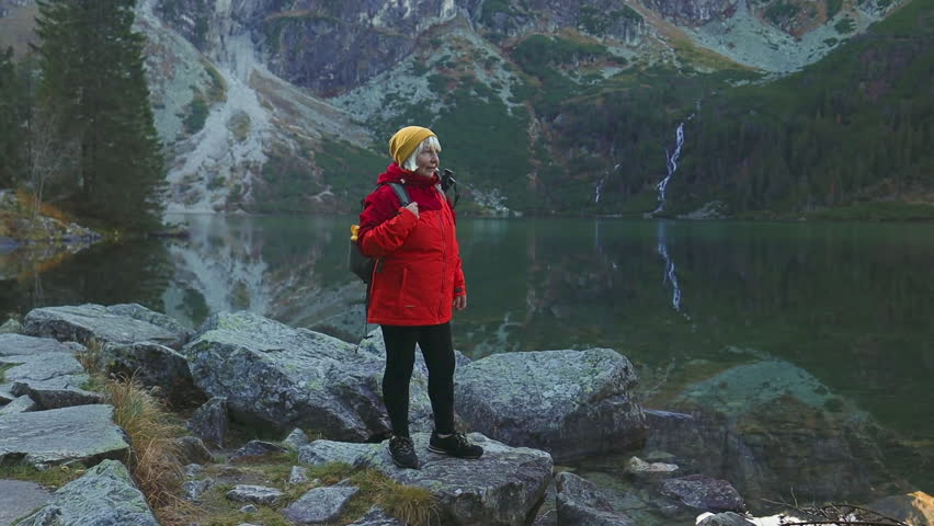 Senior woman in sport clothes with backpack trekking having fun and enjoying on a rock against a backdrop of a mountain lake in the Tatra National Park in Poland.Goal, success, freedom and achievement