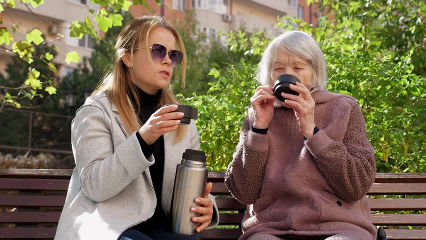 An adult woman with an old grandmother drinks hot tea sitting on a bench in an autumn city park. The granddaughter spends time with an eighty-five-year-old grandmother.