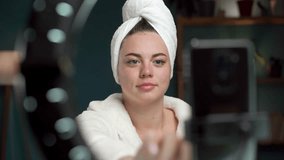 Female blogger in towel and bathrobe is showing cosmetics products while recording video and giving advices for her beauty blog, in the evening at home