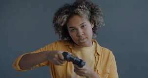 Slow motion portrait of carefree African American woman playing video game with joystick looking at camera on gray background. Entertainment and modern technology concept.