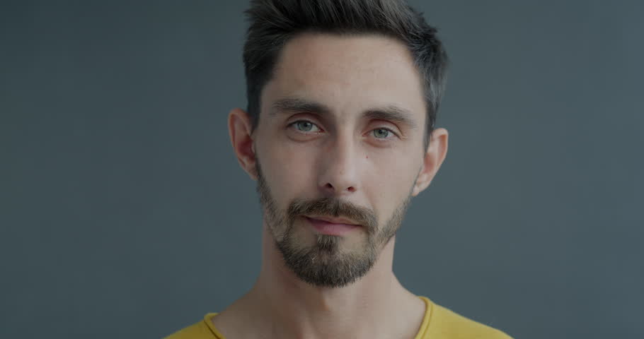 Close-up slow motion portrait of cheerful young man zipping mouth asking for silence smiling on grey color background. Secret and people concept.