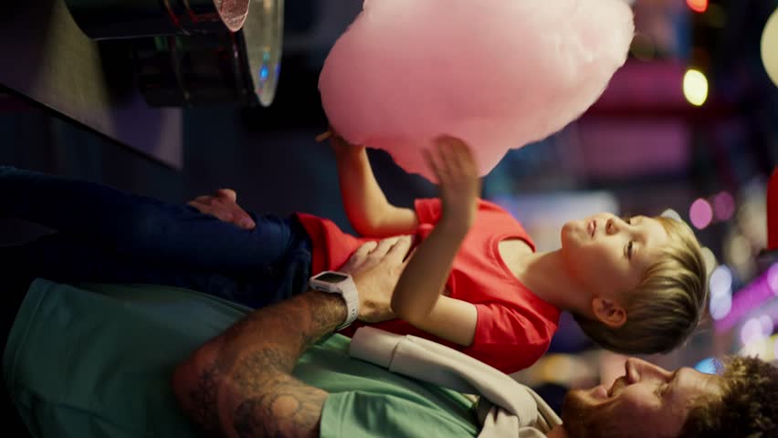 Vertical video: Little blond boy in a red T-shirt eats pink cotton candy with his dad in a Green T-shirt. Father and son going to amusement park Royalty-Free Stock Footage #1110494321