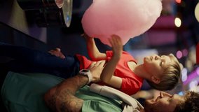 Vertical video: Little blond boy in a red T-shirt eats pink cotton candy with his dad in a Green T-shirt. Father and son going to amusement park