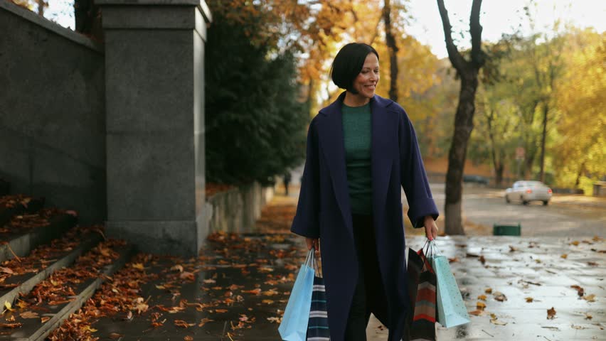 A beautiful, attractive and happy middle-aged woman is walking in the center of a city park in the autumn season in the afternoon. She is holding shopping bags in her hands Royalty-Free Stock Footage #1110504975