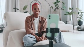 Young positive African American woman blogger draws heart with fingers recording video message on mobile phone or communicating with subscribers in live broadcast sits in stylish chair in apartment.