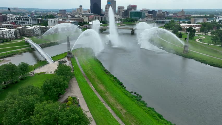 Aerial view of Dayton, Ohio: River with dramatic water fountains, pedestrian bridge, and city skyline against a cloudy sky backdrop.