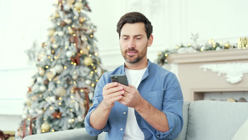 A smiling bearded man uses a smartphone sitting on sofa at home during winter New Year Christmas Xmas holidays. A handsome male is writing a greeting message, chatting with friends or shopping online Royalty-Free Stock Footage #1110512031