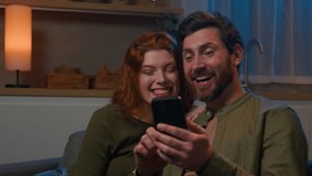 Smiling loving caucasian married couple man woman watching funny video in social media together on smartphone use mobile application in evening at home couch watch humorous movie using phone laughing