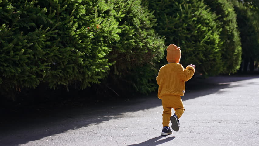Following the happy kid running by the road. Baby boy in yellow suit and cap outdoors on sunny weather.