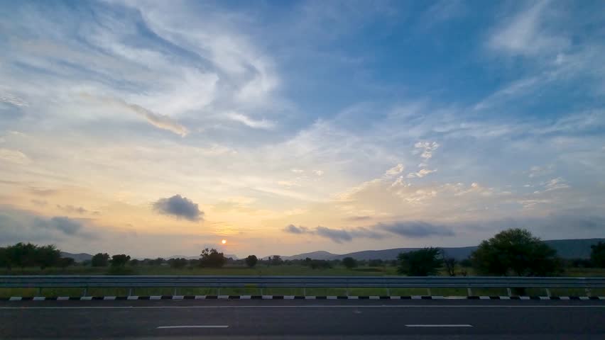 side view of road trip with signs rushing past, fluffy monsoon clouds at dusk and sunset colors in the distance showing the beauty of long drives in India