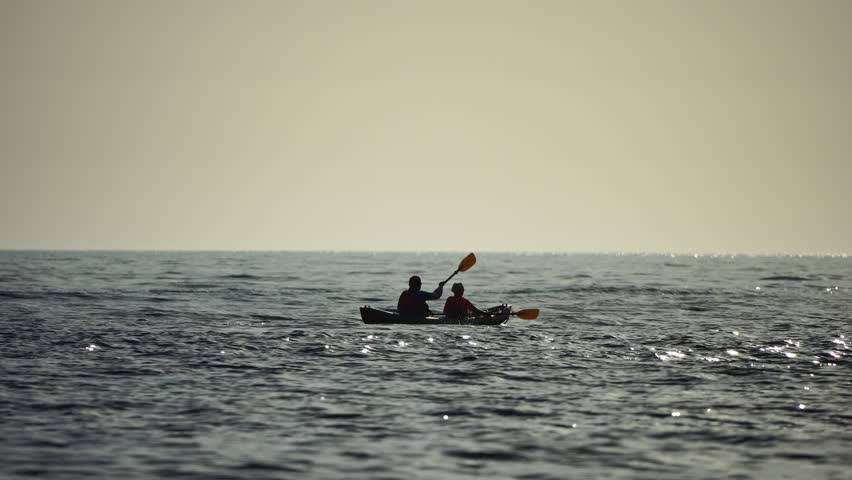 Happy couple kayaks in an inflatable kayak on the sea at sunset. Couple kanoeing in the sea near the island with mountains. People kayaking in life jackets sail. Back view