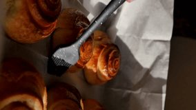 Vertical screen orientation: culinary black silicone brush brushes baked buns with sweet syrup by gently touching the baking surface on parchment. Christmas sweet cinnamon rolls