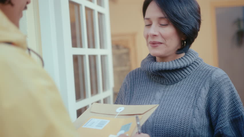 Senior Hispanic woman receiving express mail from courier, smiling and signing proof of delivery document. Over-the-shoulder shot