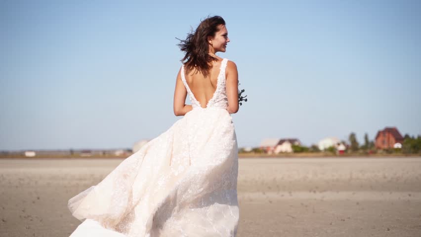 Happy young bride with bouquet of flowers going at desert landscape. Elegance lady in gorgeous wedding dress walks in nature, windy weather. Pretty woman waits for a groom after wedding ceremony. Royalty-Free Stock Footage #1110534507