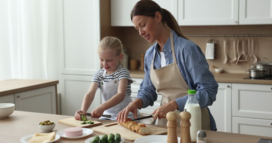 Pretty mom and little adorable kid wearing domestic aprons preparing sandwiches in kitchen, slicing bead, vegetables at table. Mother teaching little daughter girl to cook Royalty-Free Stock Footage #1110541955