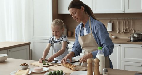 Pretty mom and little adorable kid wearing domestic aprons preparing sandwiches in kitchen, slicing bead, vegetables at table. Mother teaching little daughter girl to cook Stockvideó