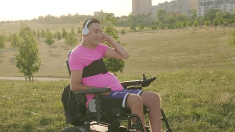 Gay man enjoys listening to music in white headphones. Sunlight illuminates man with neck tattoo sitting in wheelchair in green grassy meadow - Βίντεο στοκ