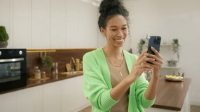 Smiling African american woman standing in modern kitchen using smart phone chatting online on video call, looking at front camera on mobile phone, enjoying modern tech. Modern real people lifestyle