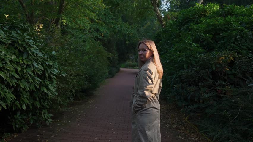 Woman in park standing sideways in light colored raincoat.