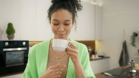 Thirsty smiling 20s multi racial woman starting new day with cup of aromatic coffee drink standing alone in modern kitchen interior. Diverse model drinking from white cup close up, people lifestyle 4K - Powered by Shutterstock - Get 15% off with code: PIKWIZARD15