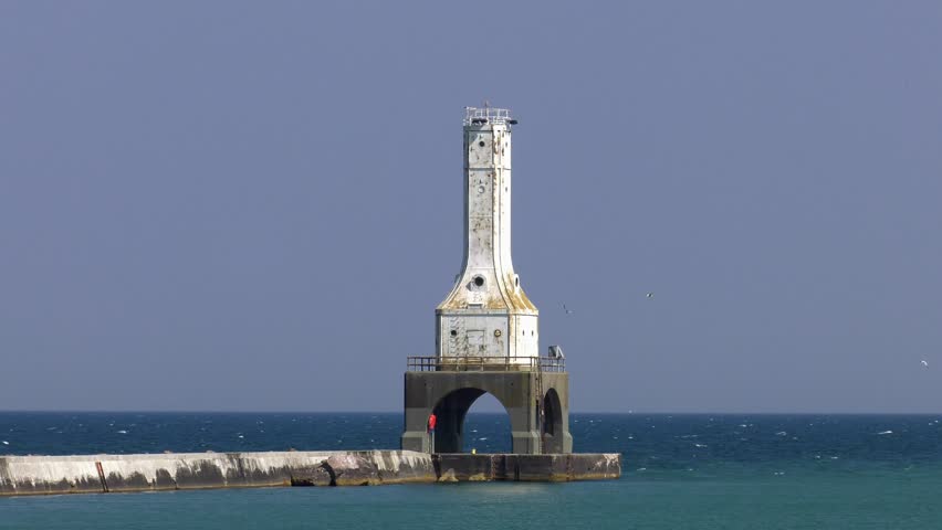 Port Washington lighthouse on Lake Michigan. Static close up shot of the lighthouse on its pier. Gulls flying around, waves breaking over the pier. Overcast day as smoke from wildfires moves in.