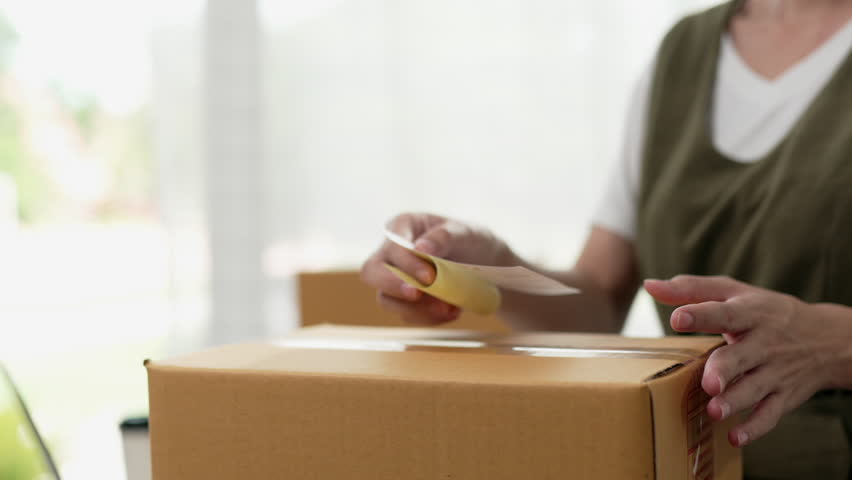 Close-up hand of a woman working adds sticker to box, packs order for client. Small business entrepreneur or owner.