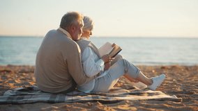 Senior couple sits on sandy sea beach hugging reading book together at sunset in evening. Happy elderly man woman family spend enjoy time together. Love couple relationship, leisure, resting concept. - Powered by Shutterstock - Get 15% off with code: PIKWIZARD15