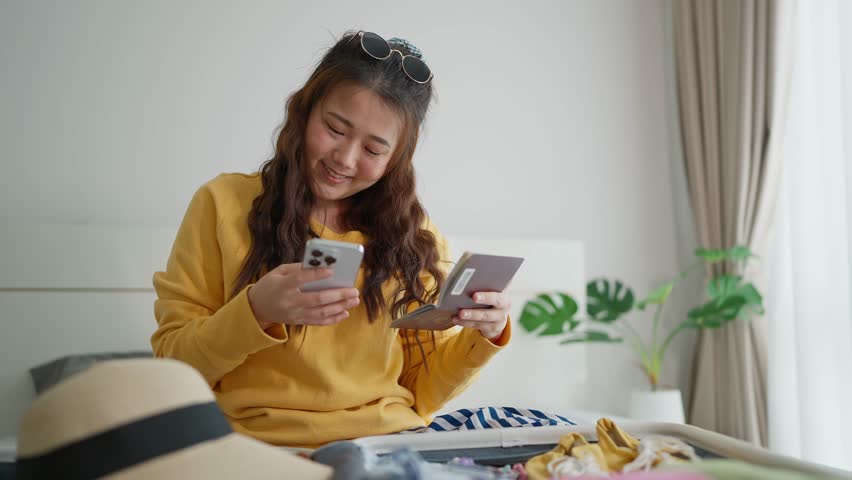 Excited young Asian woman using app booking hotel or flight on smartphone for travel while packing suitcase sitting on bed in bedroom. Booking hotel or flight before travelling on smartphone Royalty-Free Stock Footage #1110567311