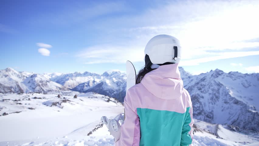 Rear view woman with snowboard watching on outdoors nature landscape with snowy hills. Girl on high top on mountains. Winter sport and wear. Enjoy rest at tour in travel. Healthy active lifestyle.