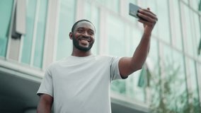 Charismatic african american man having video call on smartphone standing on city street waving hand looking at screen. Friendly guy talking on mobile phone showing thumb up. Online conversation.