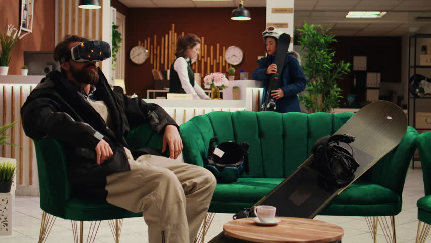 Tourist uses virtual reality technology in lobby, having fun with vr interactive headset while he wears winter suit. Guest having helmet and snowboard next to him, prepare to go sliding.