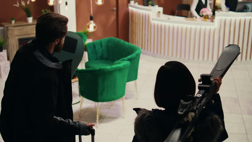 Active people enter alpine resort lobby, approaching reception staff to see room reservation while carrying skiing equipment. Young couple at mountain hotel preparing for winter sport activity.