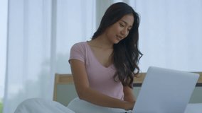 Happy professional Asian businesswoman using laptop sitting on bed at home. Smiling girl sitting, relaxing, video calling, talking about online work or study class. Take notes in a notebook.