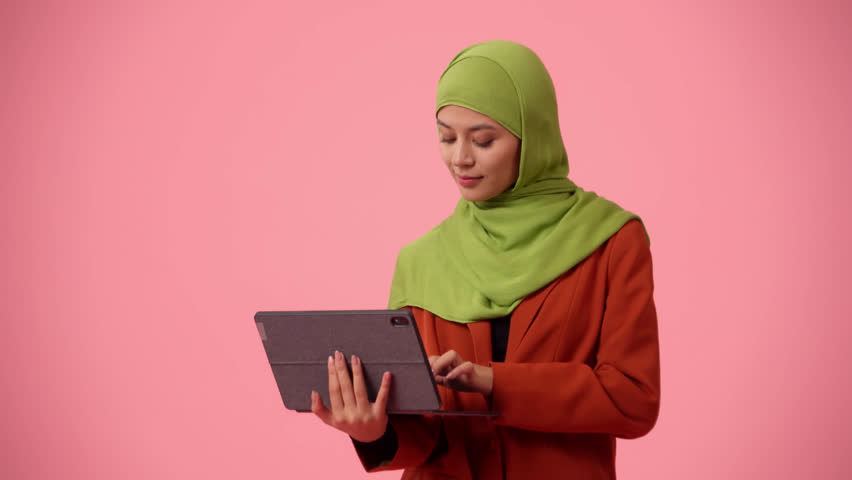 Medium-sized isolated video capturing an attractive young woman wearing a hijab, veil. She is holding a tablet, laptop, excited about great news, succesful deal.