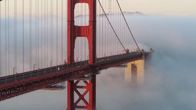 Cars driving by red bridge covered in scenic white clouds. Karl the Fog drifting below red bridge main landmark of San Francisco slow motion California USA. Aerial video cinematic Golden Gate Bridge