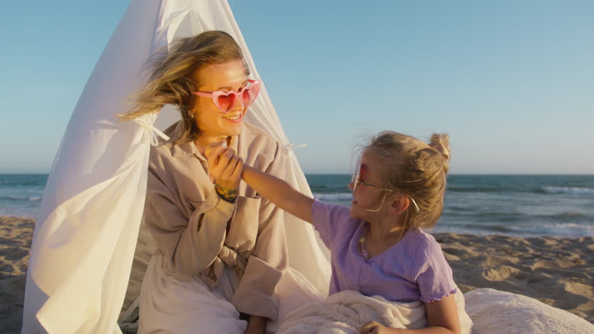 Beautiful mom gives laughing daughter high five. Young woman with girl sitting in canvas tent on ocean beach, enjoying summer vacation trip. Mother and smiling daughter having best fun time together Royalty-Free Stock Footage #1110594147