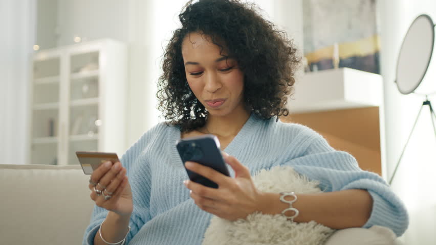 Smiling multi racial woman holding smartphone and credit card sitting on couch modern home. Happy diverse female shopper using instant easy mobile payments, customer making purchase online banking app Royalty-Free Stock Footage #1110594205
