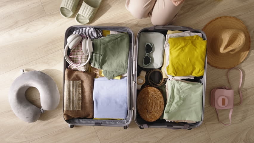 Packing suitcase top view. Efficiently packing for weekend getaway. Neatly folding vacation essentials. Unknown woman packing suitcase for trip to beach. Yellow suitcase, straw hat, flip flops Royalty-Free Stock Footage #1110598559