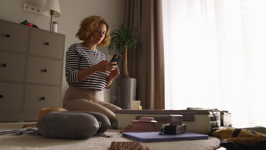 Woman packing suitcase. Woman swiftly packs clothes in suitcase. Lady sits on floor, prepares luggage for trip. Deliberate and calm, organizes belongings for journey. Leisurely packing for travel Royalty-Free Stock Footage #1110598579