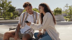Happy young multiracial couple in casual clothing using mobile phone at the skatepark