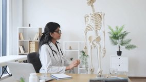 Side view of indian woman sitting at table with personal laptop near model of skeleton in personal cabinet. Brunette female doctor touching plastic bones in hospital and conducting video call.