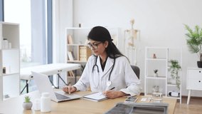Concentrated female doctor with stethoscope sitting at table and writing prescription while using modern laptop. Indian nurse looking at information about medicine and taking notes in medical office. - Powered by Shutterstock - Get 15% off with code: PIKWIZARD15