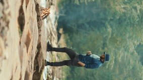 VERTICAL VIDEO: Tourist man with a mug in his hands drinks coffee or tea while standing on a stone in the mountains near a campfire. The concept of tourism and recreation in the mountains