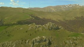 High mountains are rocky, covered with green grass and forest in clear sunny weather. Aerial view, drone video, flying around a large stone block with a man sitting on it. Pushkin Pass Armenia.