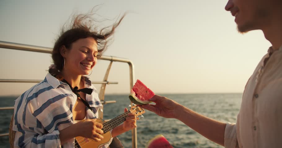 The guy invites the girl to eat a piece of watermelon at a picnic on the pier near the sea. The girl bites the watermelon and rejoices. Happy couple on a date near the sea on the pier Royalty-Free Stock Footage #1110606695