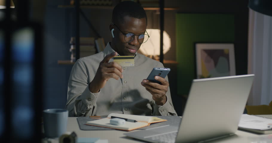 African American man using smartphone holding credit card shopping online making payment at night at home. Finance and commerce concept. Royalty-Free Stock Footage #1110608511